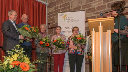 Mehrere Vereinsmitglieder des Diakonieverein Werratal e.V. stehen auf der Bühne mit Blumensträußen in den Händen beim Zusammenschluss von Diakonie Werratal und Immanuel Diakonie Südthüringen im Kressehof im Meininger Ortsteil Walldorf Mehrere Vereinsmitglieder des Diakonieverein Werratal e.V. stehen auf der Bühne mit Blumensträußen in den Händen beim Zusammenschluss von Diakonie Werratal und Immanuel Diakonie Südthüringen im Kressehof im Meininger Ortsteil Walldorf